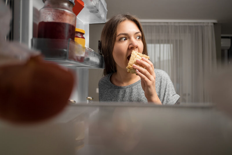 A young woman is eating directly from the fridge late at night, the cold food is making her stomach feel cold.