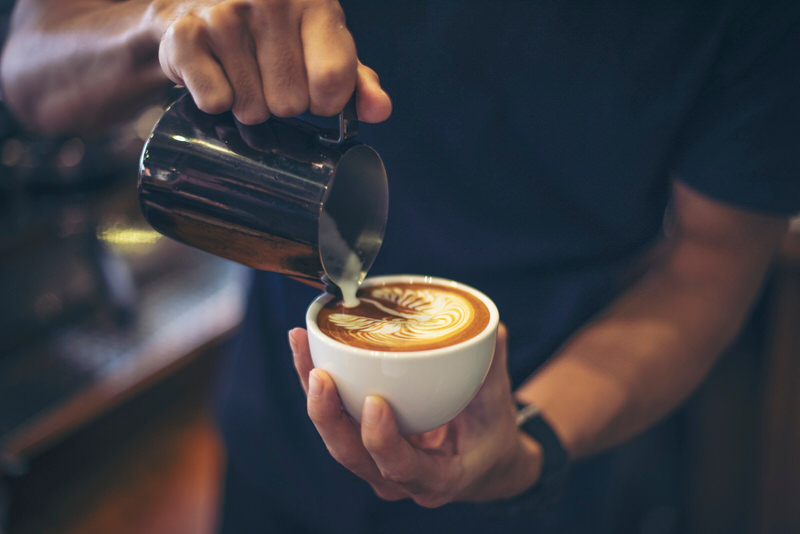 A barista is making a cappuccino with milk and sweetner.