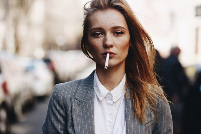 A young woman in work attire is smoking outside during her work break.