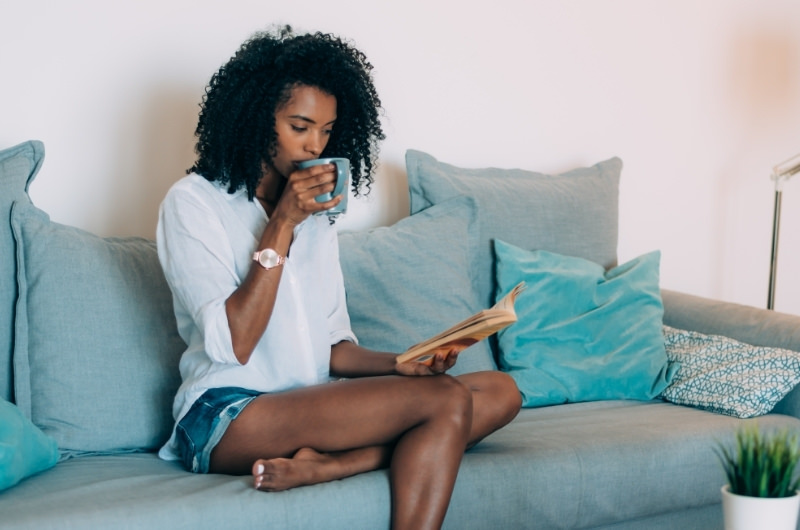 A young woman is sitting on her sofa at home, reading a book, and drinking black coffee.