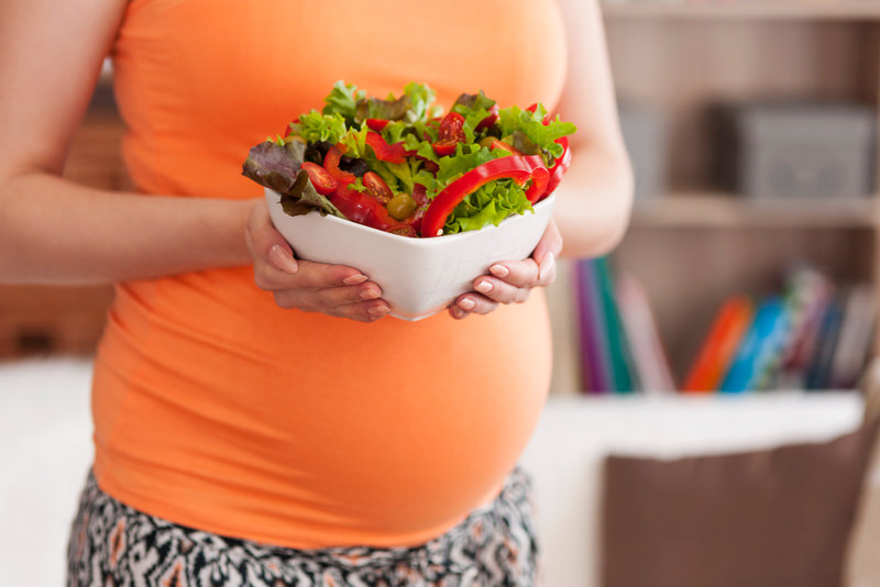 A young pregnant woman is holding a large salad bowl, to eat as an alternative to other convenient but less healthy options.