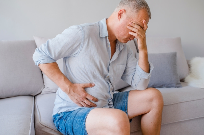 A middle aged man is sitting on the sofa clenching his right side of his stomach because of sharp pain, a possible sign of Appendicitis.