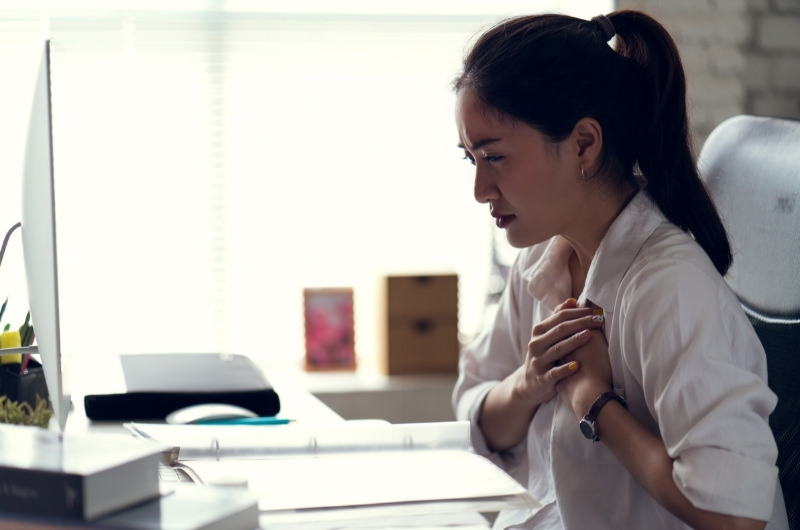 A young woman is in noticeable pain as she's holding her chest while sitting at her office desk.