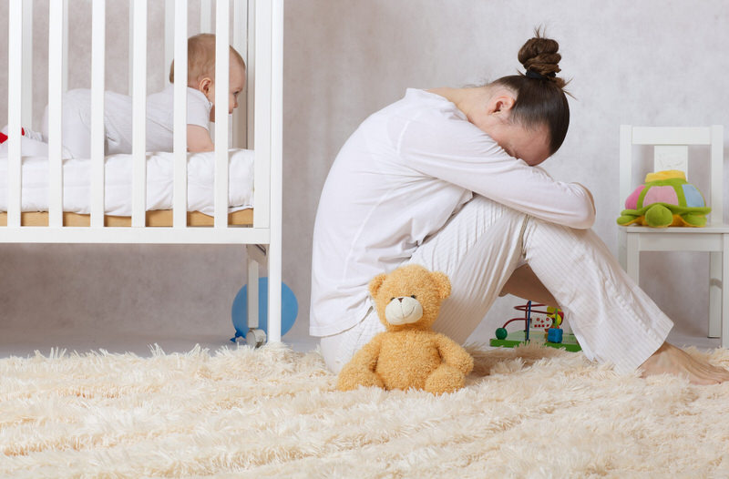 A new mom who's experiencing postpartum depression is sitting next to her baby's crib tired and unhappy.
