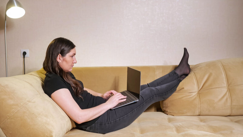 A woman who's traveling is working in her hotel room with her legs raised, to allow her ankles swelling to go down.