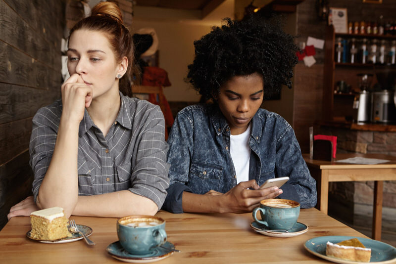 A young woman is at a coffee shop with her friend, but is not conversing with her friend, she has a hard time making small talk.