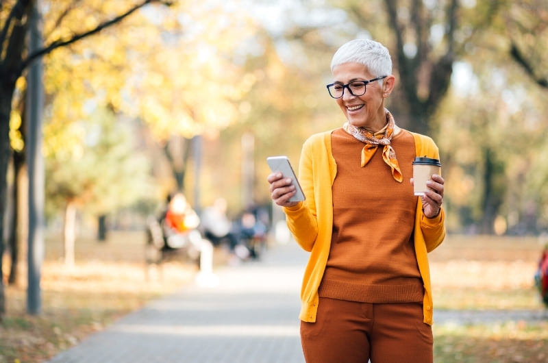 An older woman is holding coffee and walking in the nearby park to avoid sitting for too long and prevent varicose veins.