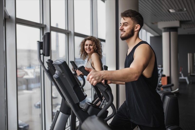 A young man and woman are using the elliptical at the gym next to each other.
