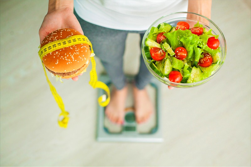 A woman is standing on a scale while holding a salad in one hand and a burger in another. She's struggling to balance her weight loss goals with her food cravings.