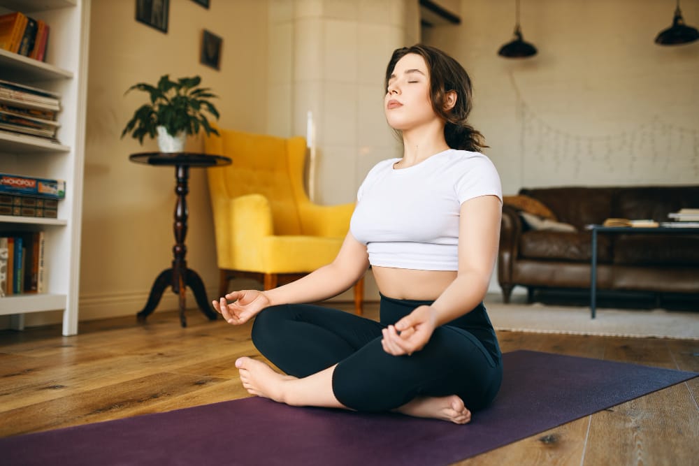 A young woman is sitting on her yoga mat meditating and doing a breathing exercise to help ease her headache