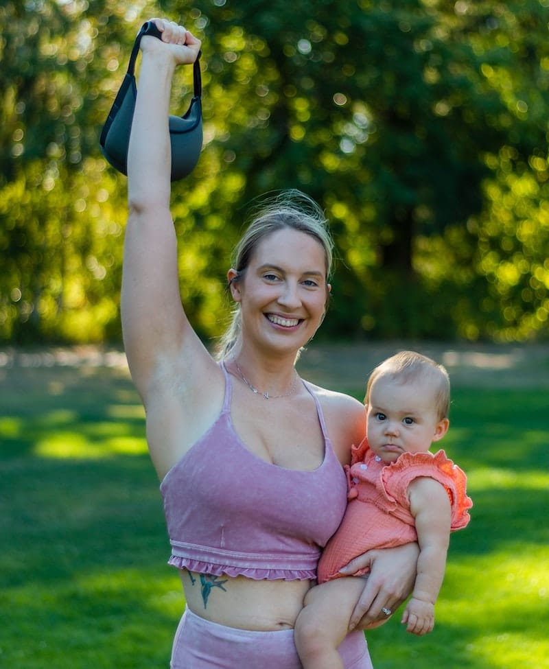 A young mom is holding her baby and a kettlebell, which she will use soon during her exercise session.
