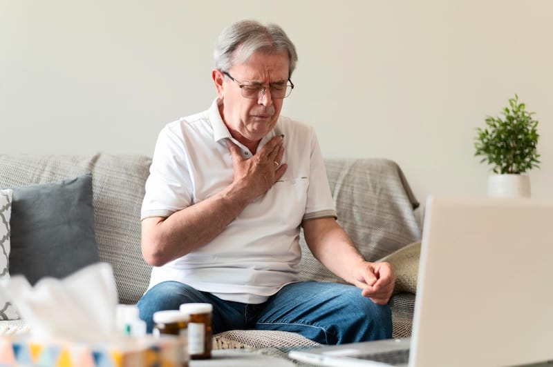 An older man sits on his sofa, clenching his heart in pain. He may be having a heart attack.