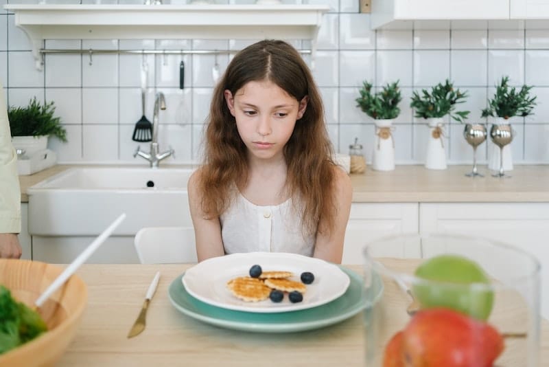 A young girl with early satiety is not able to eat the blueberry pancake breakfast she has in front of her