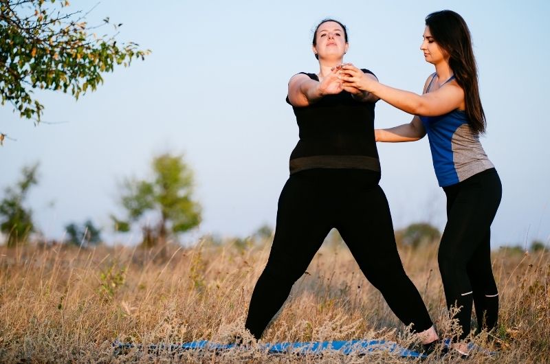 A woman is exercising outside on a mat with her personal trainer guiding her