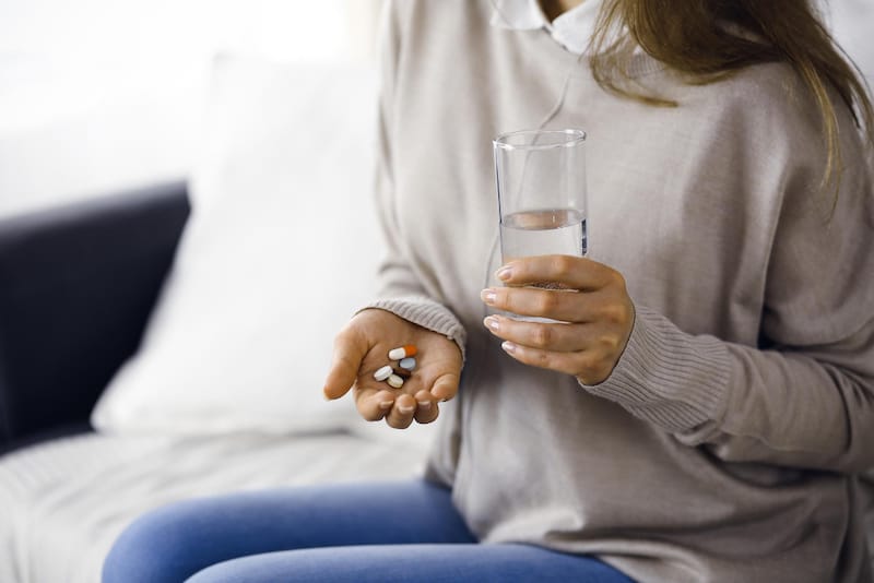 A woman is holding 5 different medicines in her hand while holding a glass of water in the other hand