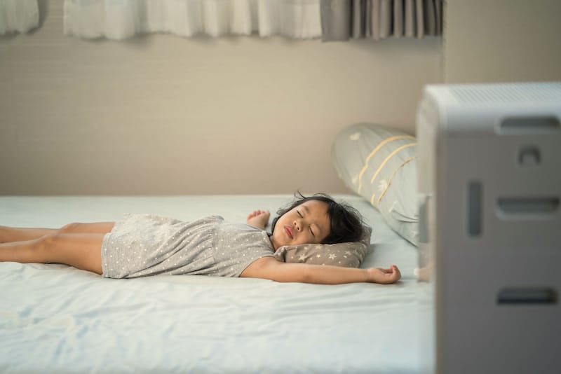 A young girl is sleeping on her bed with the air purifier turned on next to her
