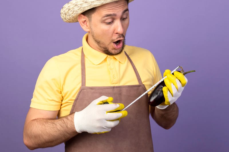 A man is holding an eggplant and measuring it with a tape ruler as an example of how age can affect the size of a penis over time.