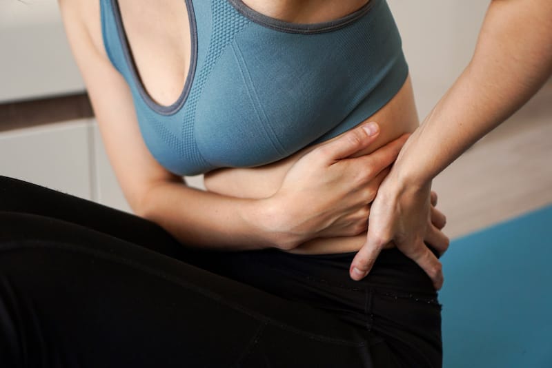 A woman is sitting down and holding on to the left side of her waist, as it's causing her a lot of pain.