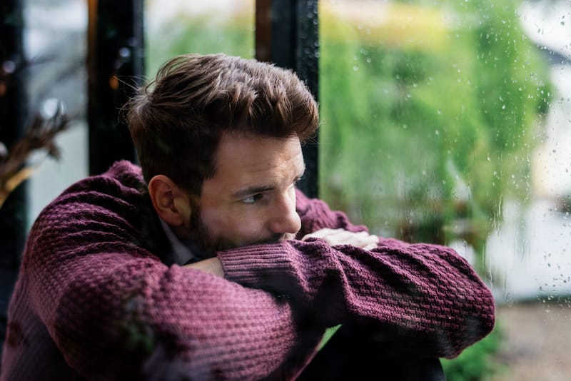 A young man who's experiencing seasonal affective disorder is sitting in his home looking outside at the rainy day