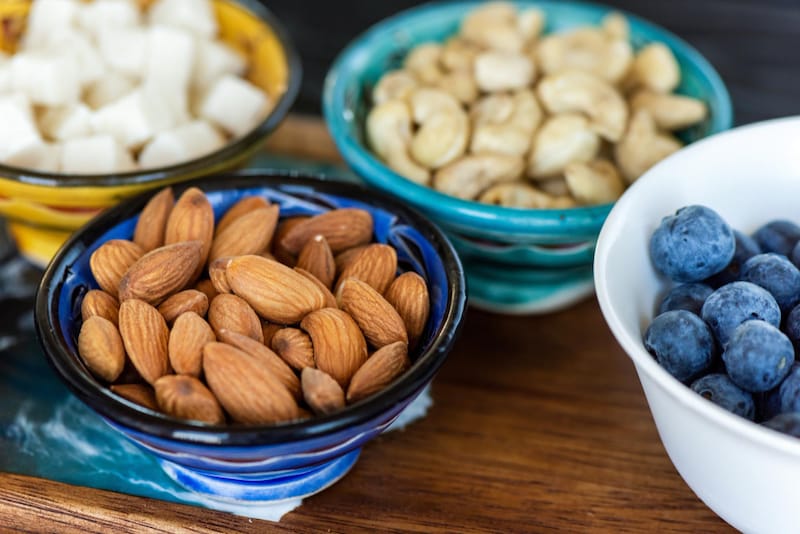 Various bowls of healthy snacks are shown, including almonds, cashews, and blueberries.