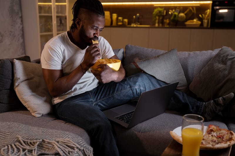 A man is bored eating late at night while watching something on his laptop