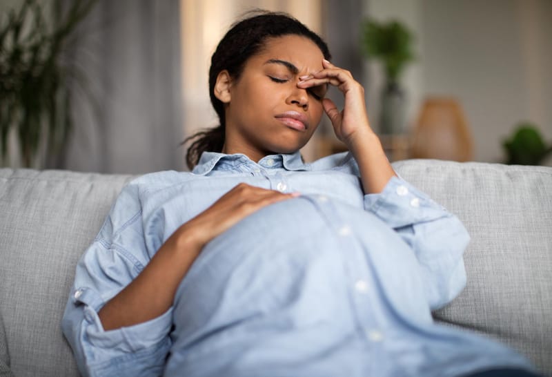 A pregnant woman is sitting down with her eyes closed as she is dealing with her headache