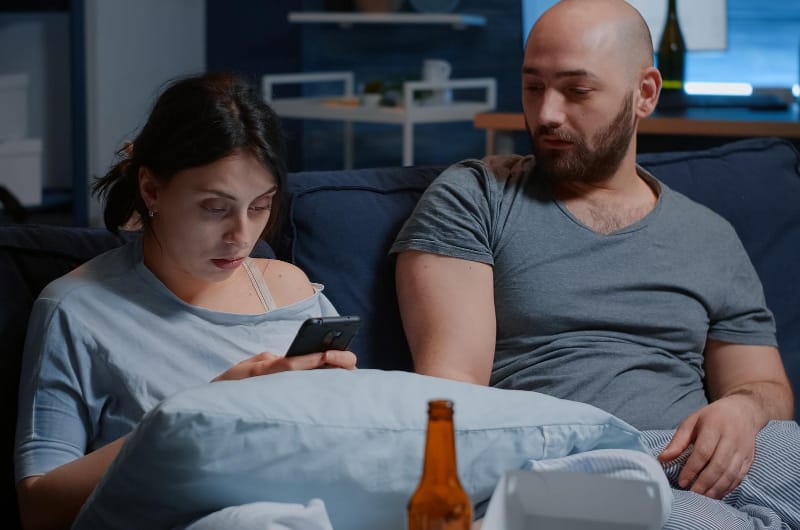 A man is sitting with his girlfriend on the sofa, trying to help her cope with her anxiety while she tries to drown out the world by using her phone.