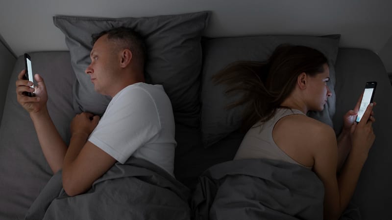 A young couple is in bed looking at their phones in separate direction, as they're not getting along at the moment.