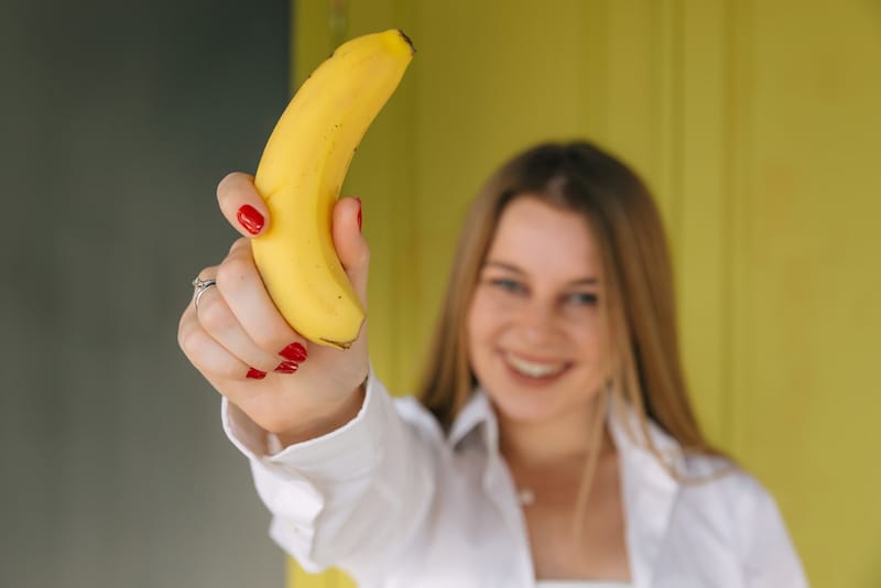A woman is smiling and holding up a banana that she's about to eat