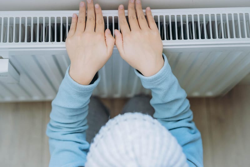 A young boy is placing his hands on an indoor heater to warm his cold hands, a symptom of his arterial thoracic outlet syndrome