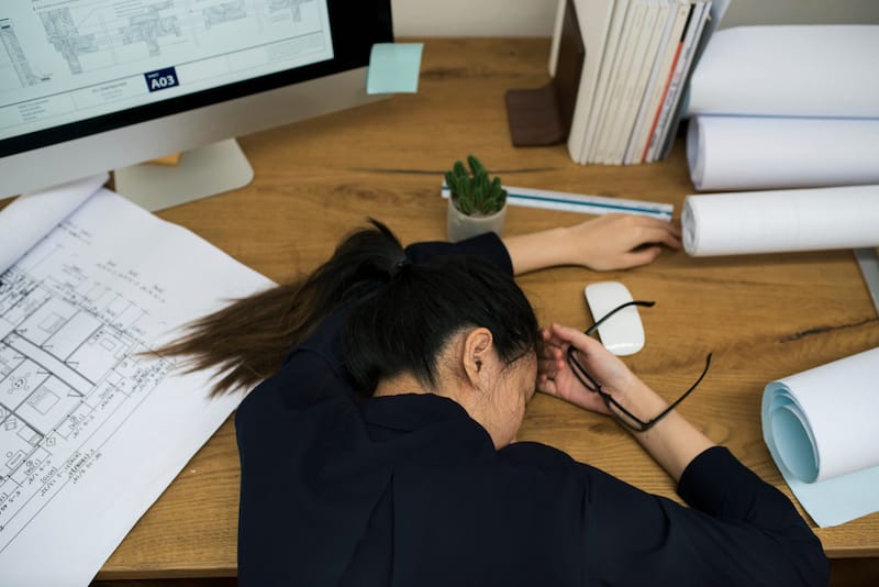 A young woman looks noticeably anxious as she puts her head down on her office desk, feeling tired.