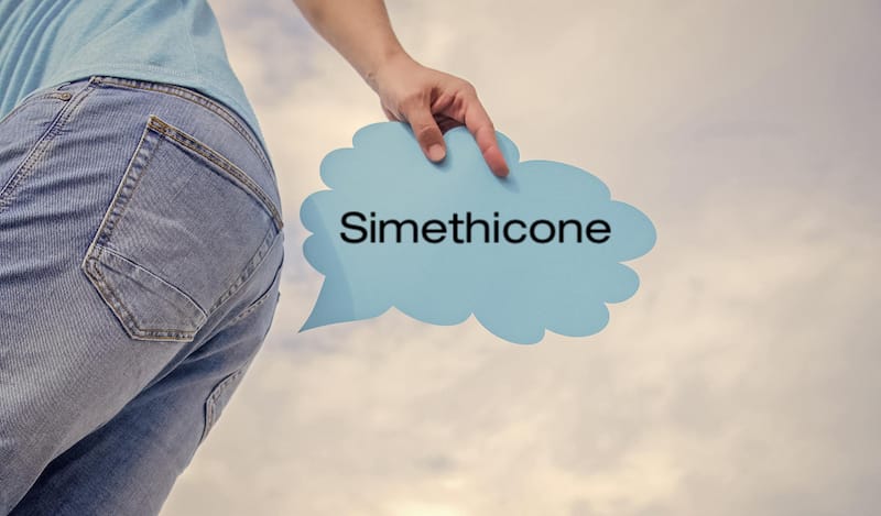 A man has a sign that says "Simethicone" and is pointing it at his butt to represent excess gas in the body coming out as a result of taking the medicine Simethicone.