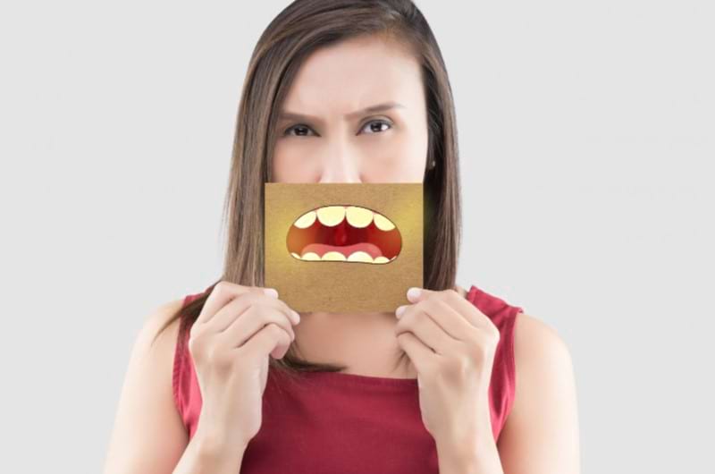 Asian woman holding a paper with yellow teeth cartoon drawn on them hiding her mouth.