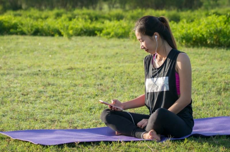 An Asian woman is sitting on a yoga mat in the middle of a green ground and is listening to music, trying to relax to cope with her anxiety.