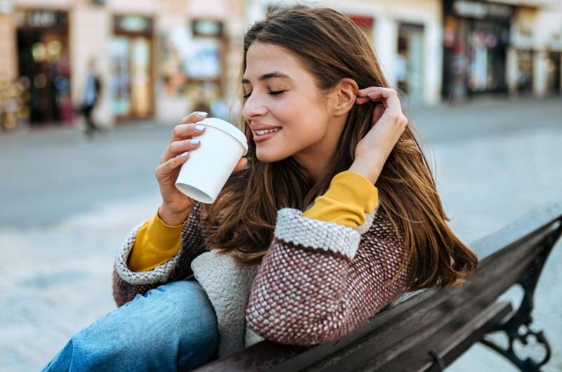 A young woman sitting on a bench outside sipping on a cup of coffee is a potential cause behind yellow teeth.