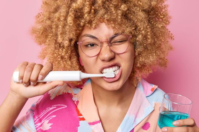 A woman with curly hair is brushing her teeth aggressively while holding a mouthwash in a glass in her other hand, focussed on whitening her yellow teeth.