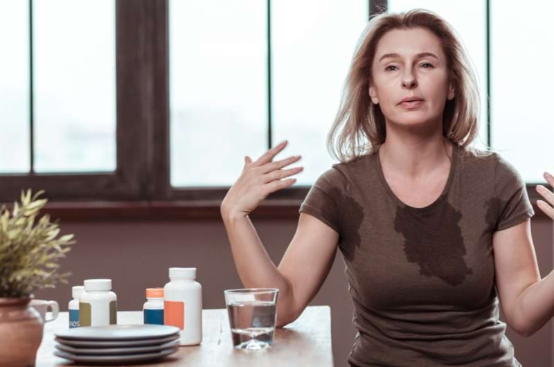 A middle-aged woman is sweating while suffering from hormonal issues with several medicines kept on the table beside her.