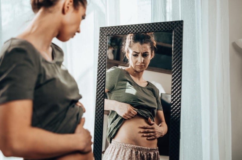 A young woman is frowning, looking at herself in the mirror after her fibroid removal surgery, unable to lose belly fat.