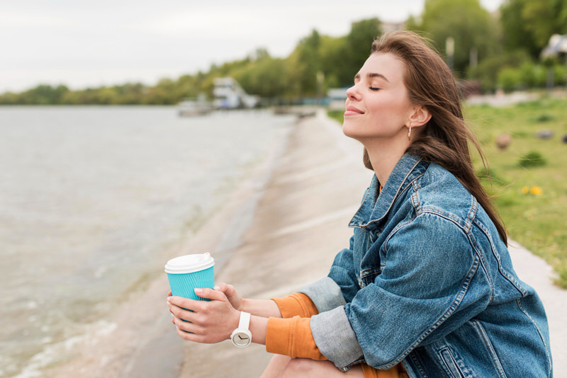 A woman is happy as she's sitting by the beach, enjoying the breeze and drinking a cup of coffee