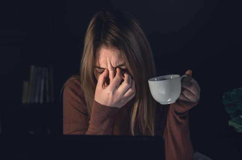 A young woman is awake at night drinking coffee while experiencing a headache, unable to have a good night's sleep.