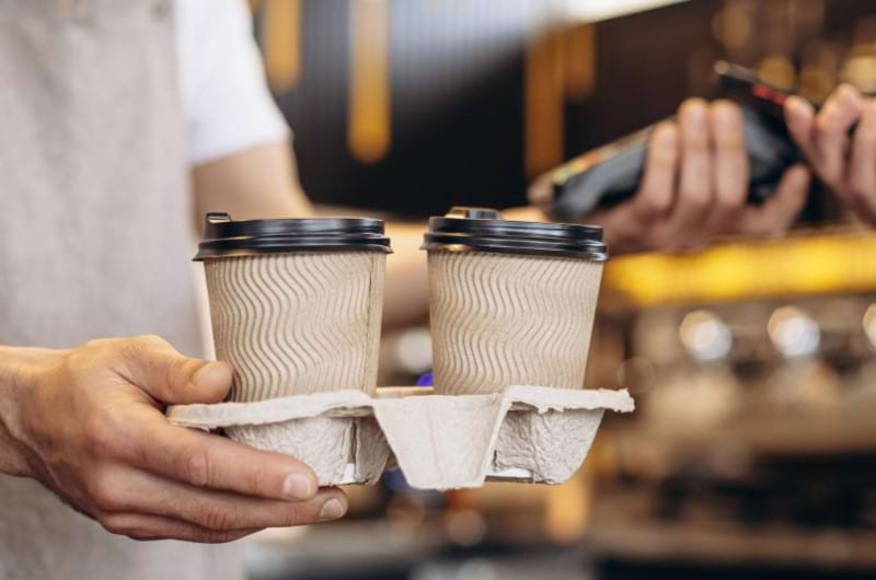 A male barista is holding a tray with two coffees and doing payment with other hand.