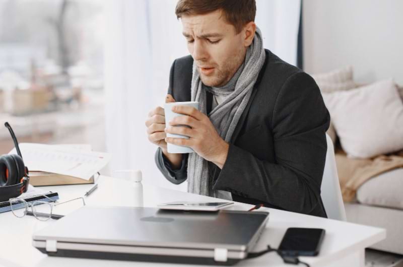 A sick man is sitting by the table shivering with a cup of coffee in hands and ready to work.