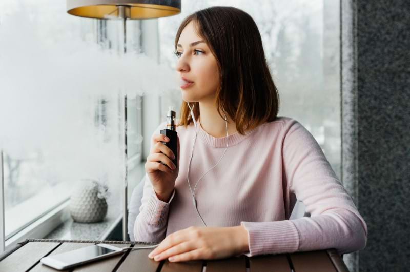 A woman sitting down and vaping as she looks outside the window. 