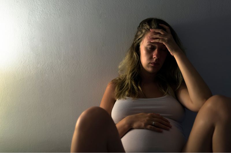 A pregnant woman is sitting by herself on the ground looking noticeably sad and anxious
