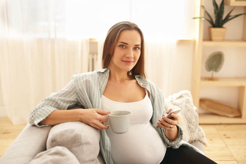 A pregnant woman is sitting down on her sofa having a cup of coffee in the morning