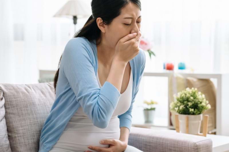 A nauseated pregnant woman is sitting on sofa struggling with not to throw up.
