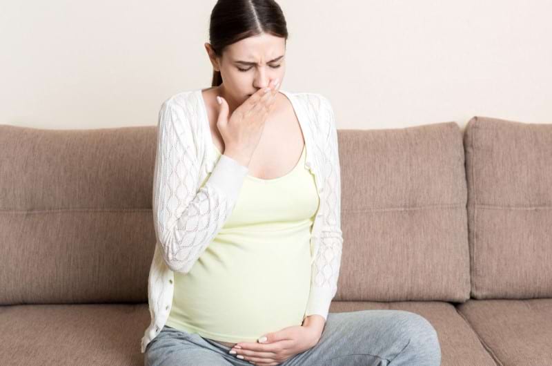 A pregnant woman sitting on the sofa is feeling nauseated due to pregnancy hormones.