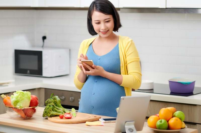 A young pregnant woman is standing in her kitchen noting down healthy recipes in her notepad while surrounded by healthy fruits and veggies. 