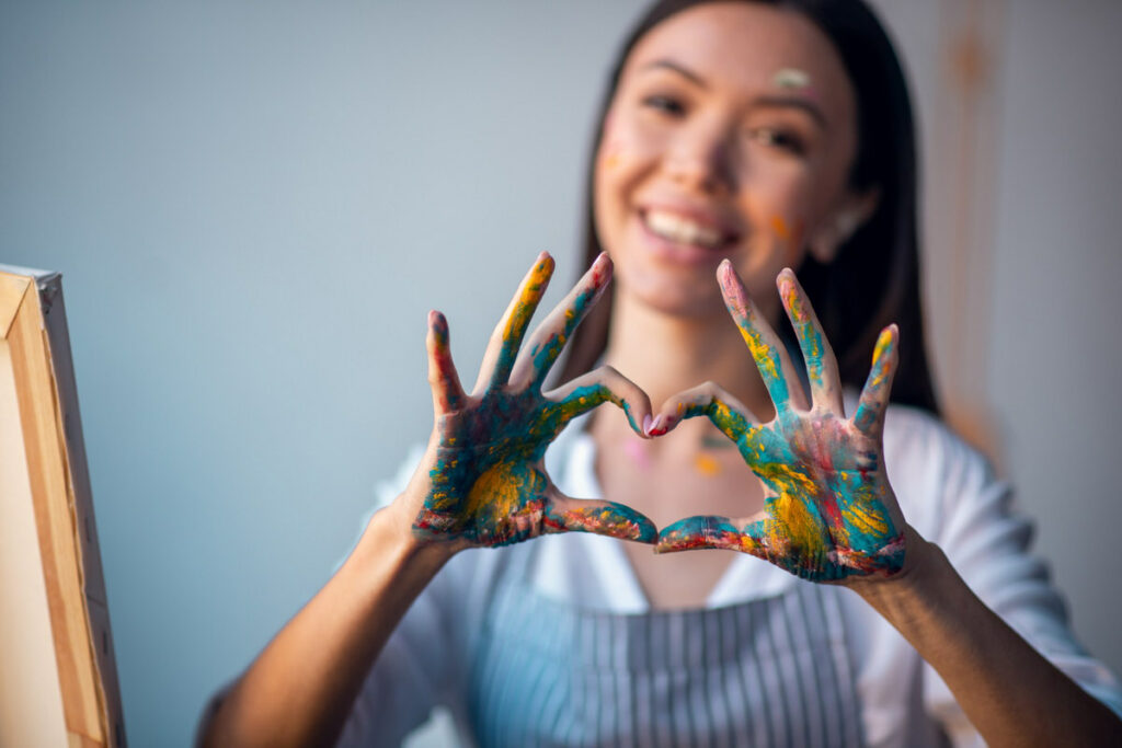 A young woman is who is painting on canvas is happy and doing a heart sign with her hands to show she's happy