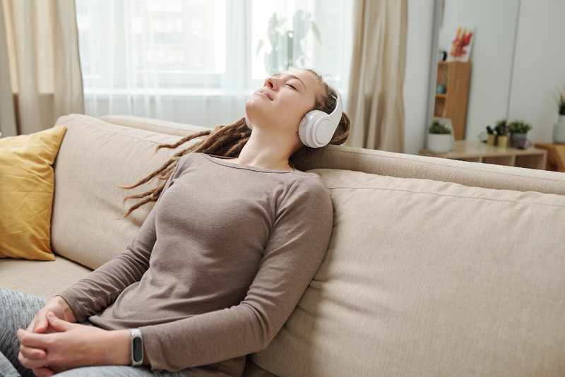 A young woman laying on the sofa with her eyes closed, listening to music while she meditates.